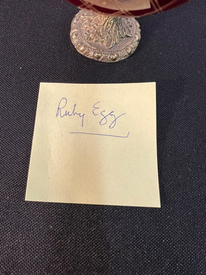 Note with 'Ruby Egg' written on it, placed under a silver coin on a dark fabric background.