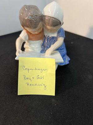 Ceramic figurine of a boy and girl reading with a yellow note on a black surface