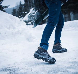 Person wearing black winter boots and blue jeans walking on a snow-covered ground with trees in the background.