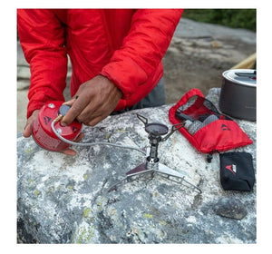 Person in red jacket using a camping stove on a rock with outdoor gear around