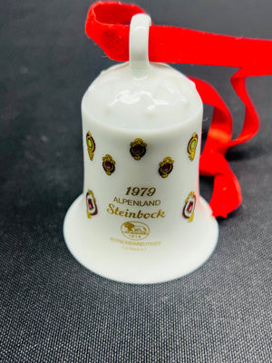 White bell-shaped ornament with gold text and logos on a red ribbon against a dark background