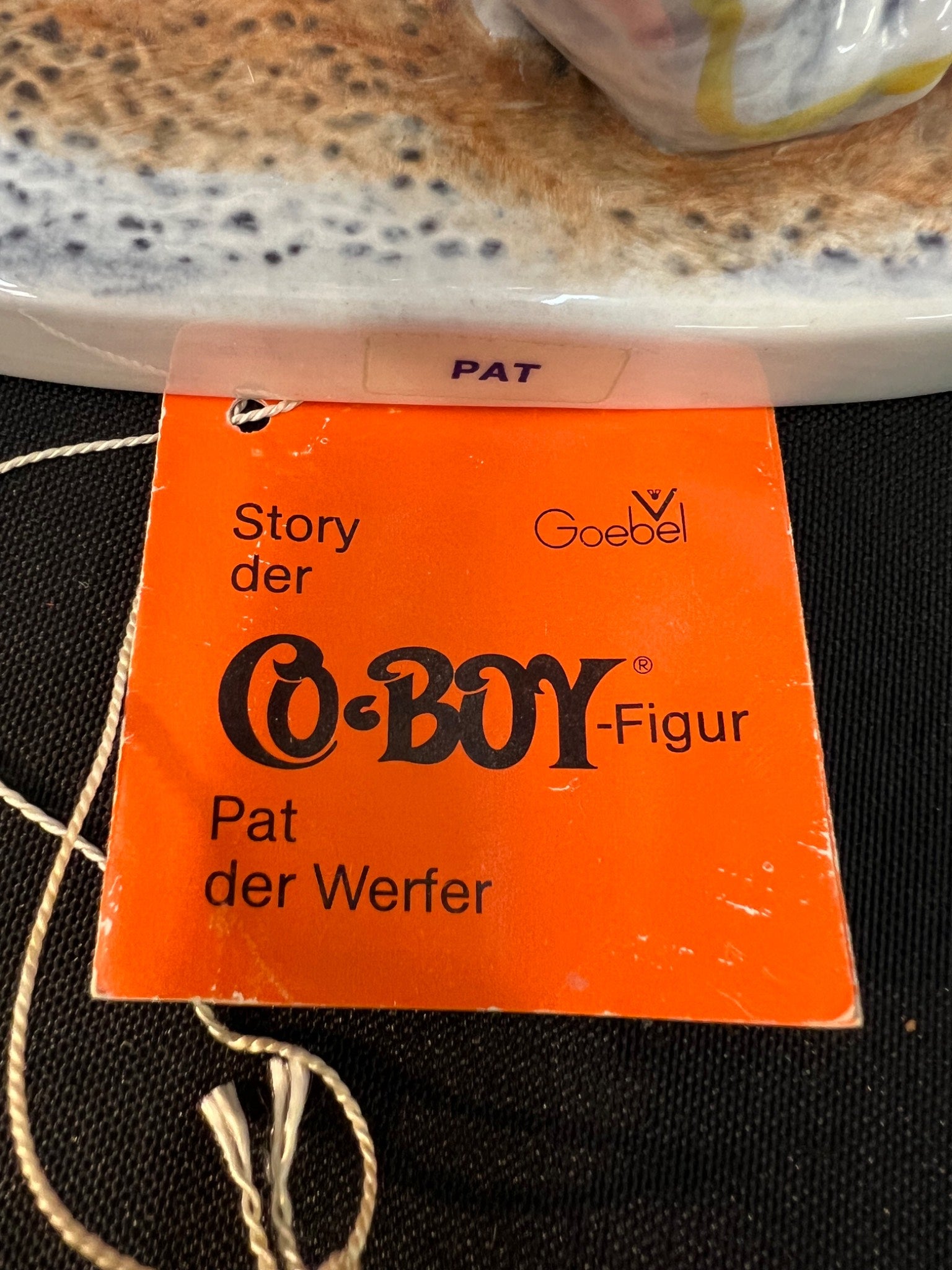 Orange tag with text about a Goebel Co-Boy figurine on a textured surface