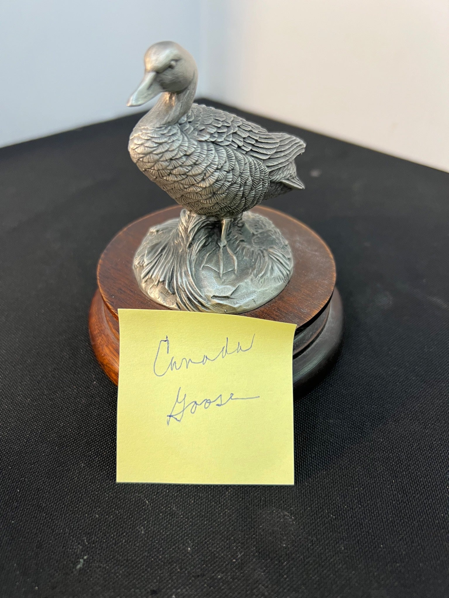 Bronze duck sculpture on a wooden base with a yellow sticky note labeled 'Canada Goose' on a black surface.