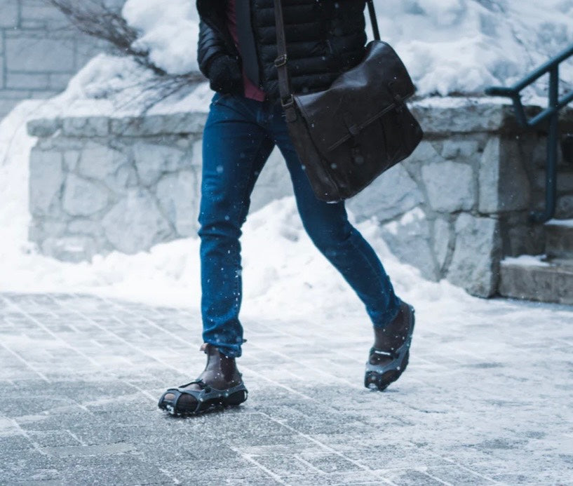 Person walking in snow wearing blue jeans and black shoes with a black bag.
