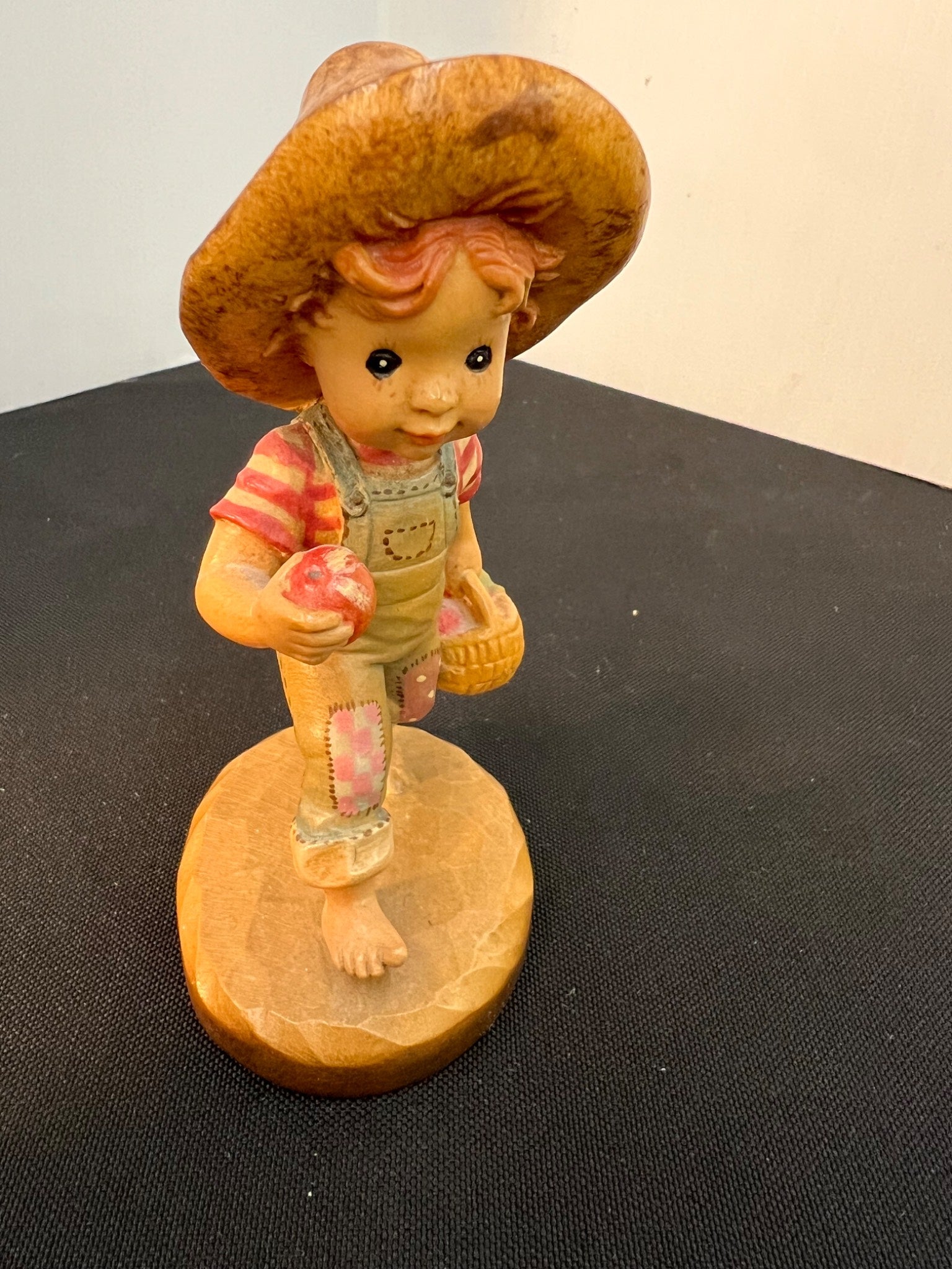 Vintage figurine of a child in overalls and hat holding an apple on a wooden base.