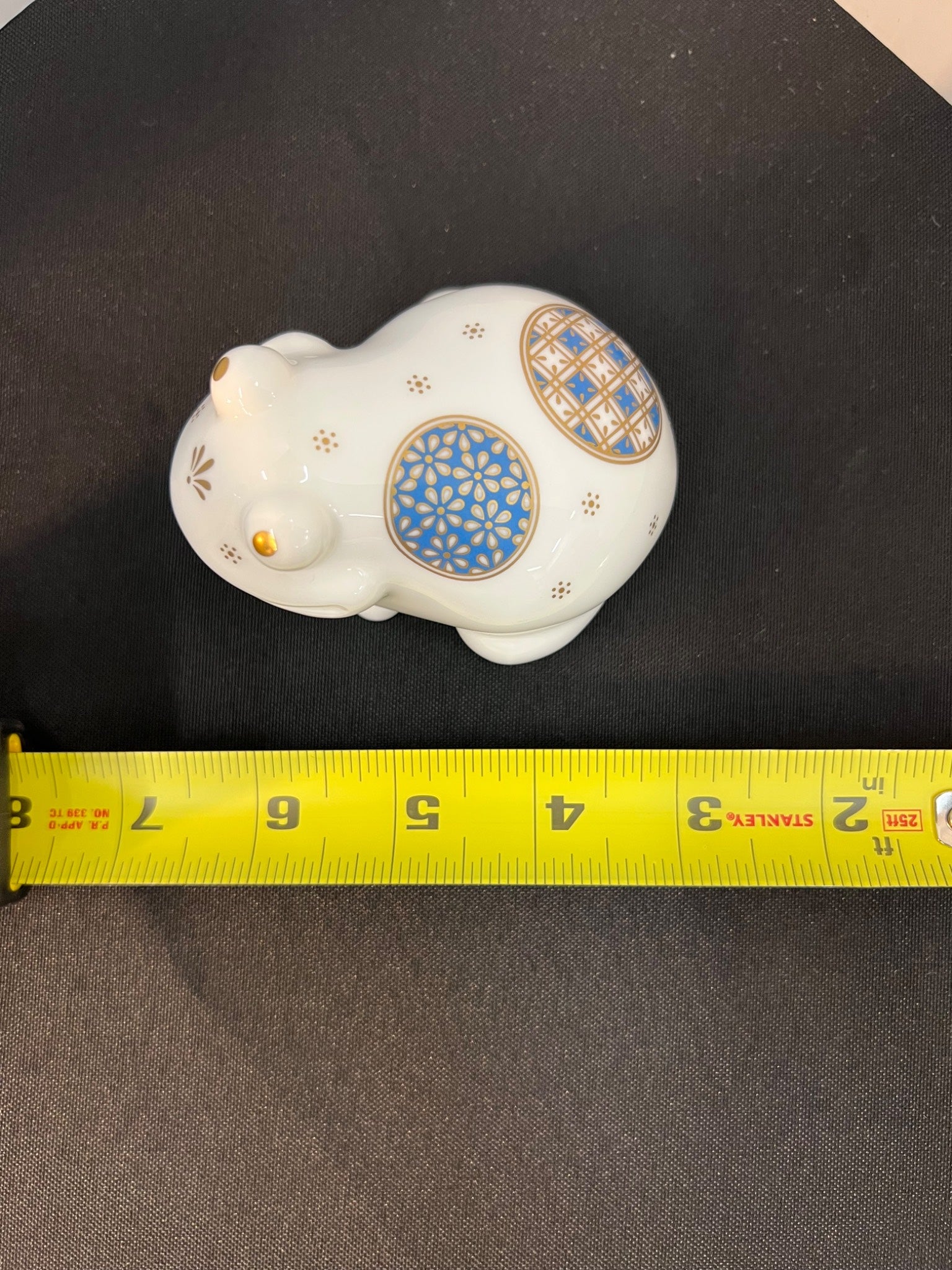 Decorative white figurine with blue and gold patterns on a black surface, measured by a yellow tape measure.