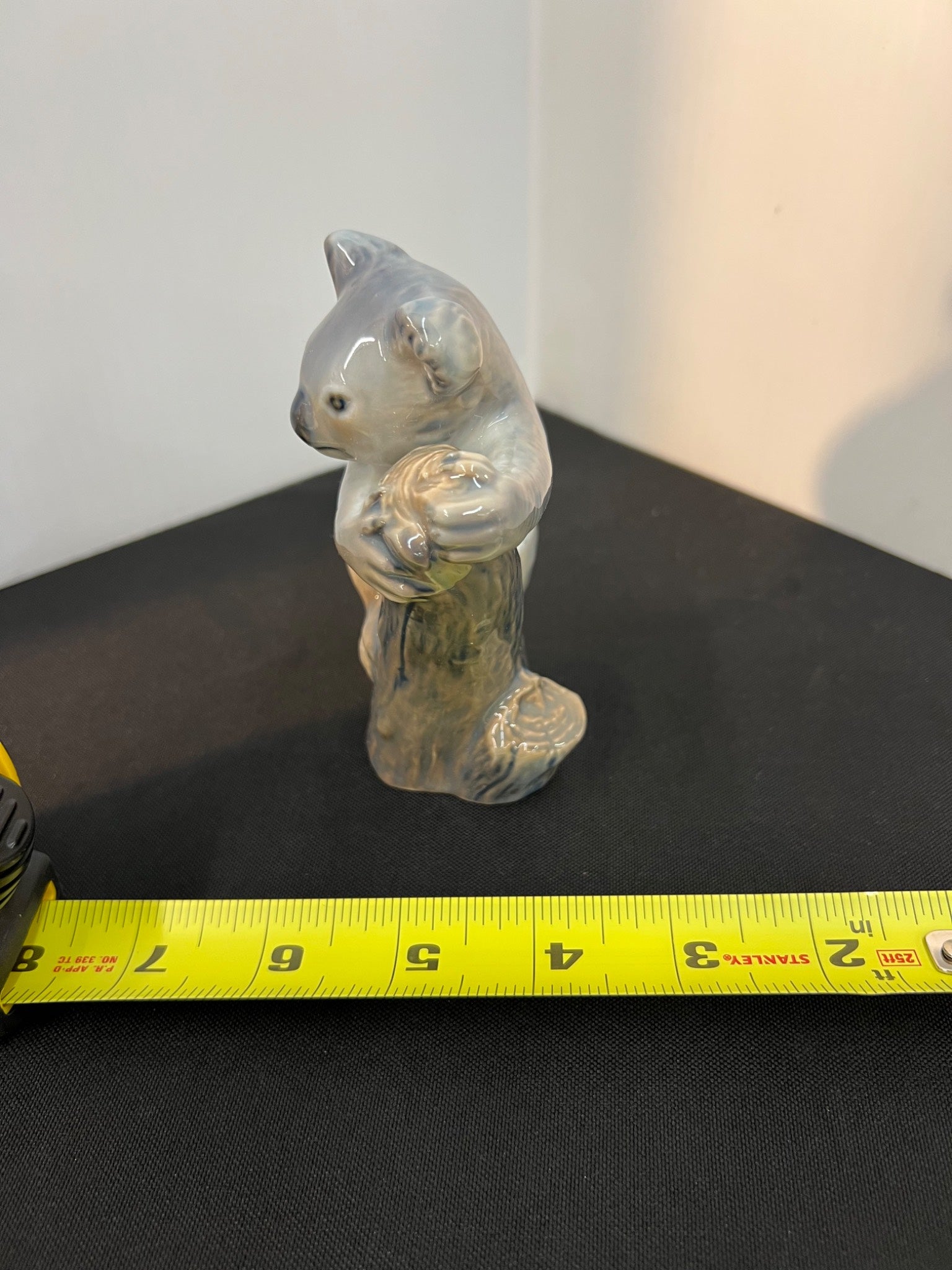 Small ceramic koala figurine on a black surface with a measuring tape for scale.