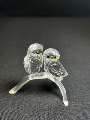 Glass sculpture of two owls on a branch against a dark background