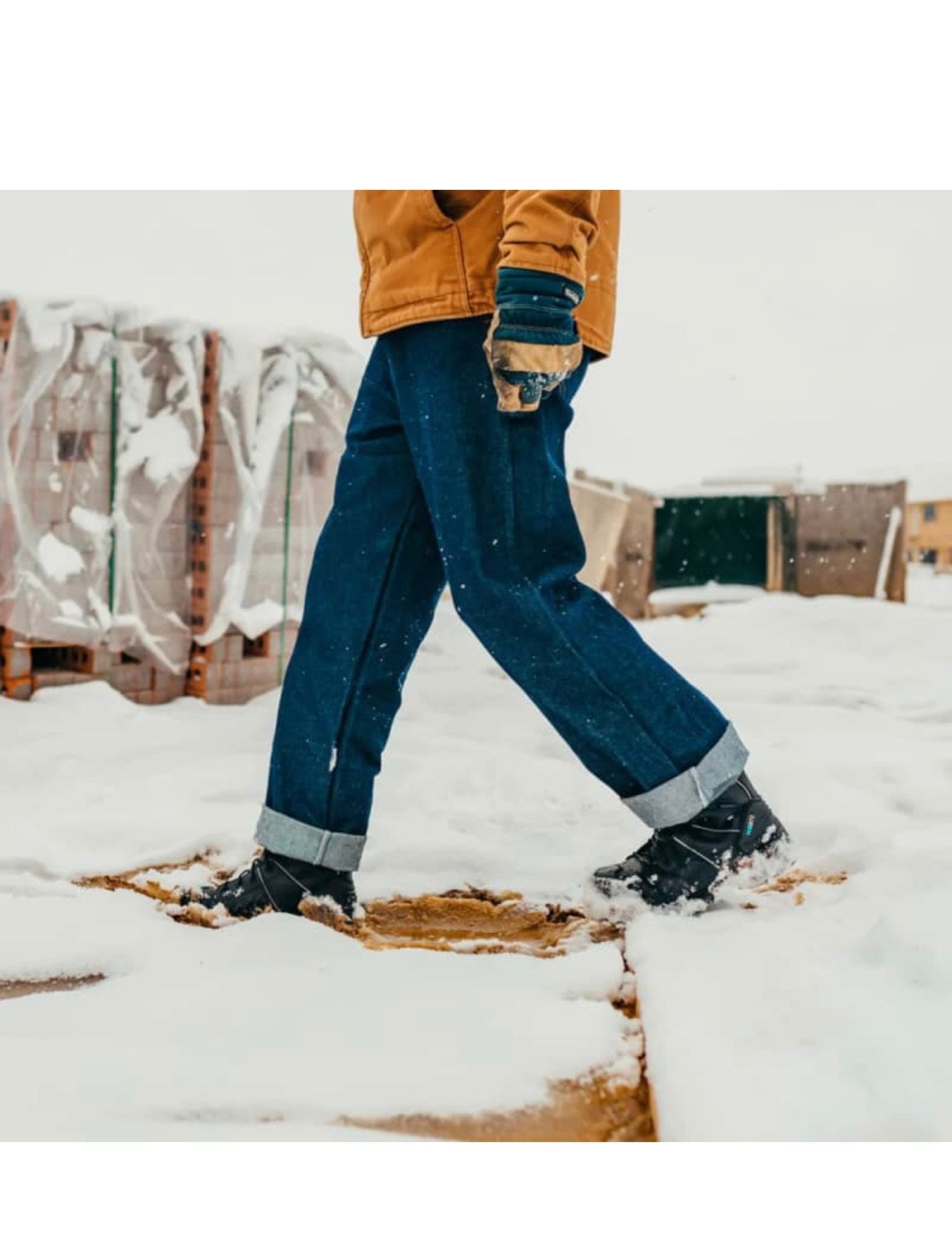 Person wearing blue jeans and black boots walking on a snowy surface with wooden pallets in the background.