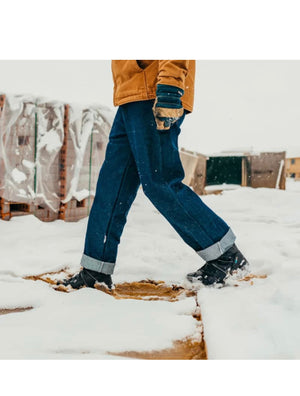 Person wearing blue jeans and black boots walking on a snowy surface with wooden pallets in the background.
