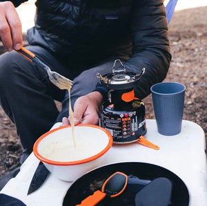 Person camping, cooking outdoors with a portable stove and utensils.