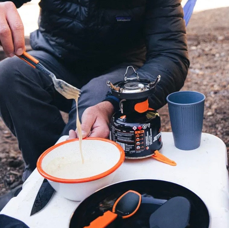 Person camping, cooking outdoors with a portable stove and utensils.