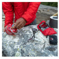 Person in red jacket using a camping stove on a rock with outdoor gear around