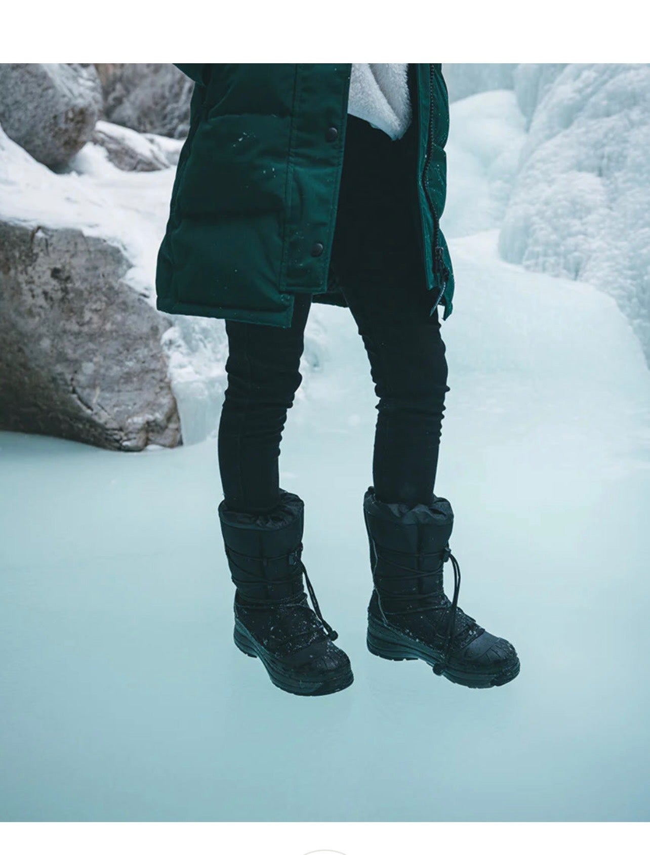 Person wearing a green coat and black boots standing on icy ground with snow and ice formations in the background.